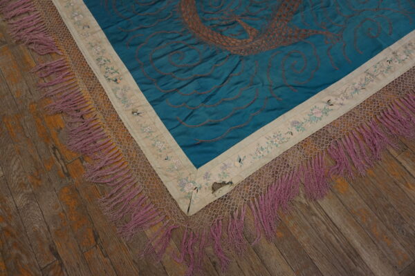 Embroidered blue rug corner featuring brown stitching, a floral border, and long mauve fringe over wood flooring.