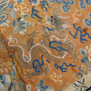 Ornate textile with metallic gold dragon embroidery, blue clouds, and orange flowers on a saffron background.