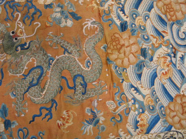 Detail of a textile with embroidered green dragon, vibrant blue waves, and large yellow flowers on a shimmering gold ground.