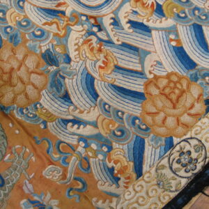 Detail of embroidered textile with blue waves, tan flowers, and a patterned border on wood flooring.