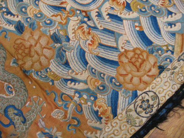 Detail of embroidered textile with blue waves, tan flowers, and a patterned border on wood flooring.