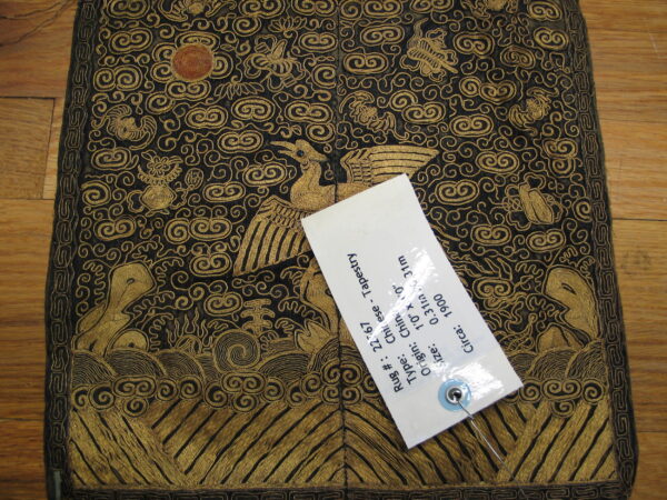 Black textile with extensive gold metallic embroidery of birds, clouds, and waves rests on a wood floor.