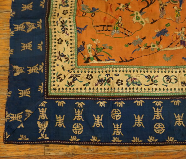 Orange embroidered textile featuring figural scenes and blue borders with gold characters, laid on wood floor.