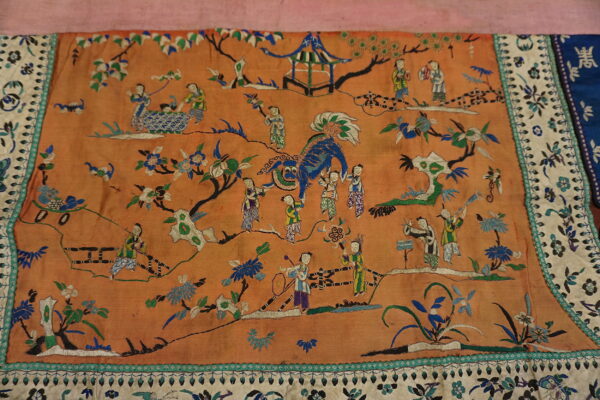 Low-pile orange textile featuring detailed embroidery of figures, a blue creature, a pavilion, and a cream floral border.