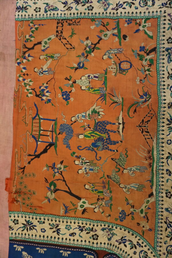 Terracotta flat textile embroidered with colorful traditional figures, trees, and ornate blue and cream multiple borders.