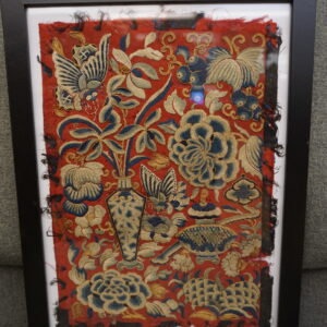 Framed red textile fragment with detailed blue and gold floral embroidery on gray upholstery.