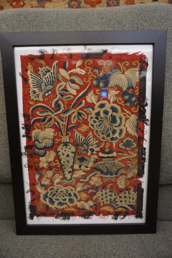 Framed red textile fragment with detailed blue and gold floral embroidery on gray upholstery.