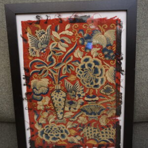 Black-framed red and blue embroidered fabric on a gray sofa, with a beige patterned rug visible in the background.