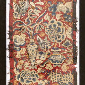 Framed red textile with dense blue and gold embroidery depicting flowers, butterflies, and a patterned vase.