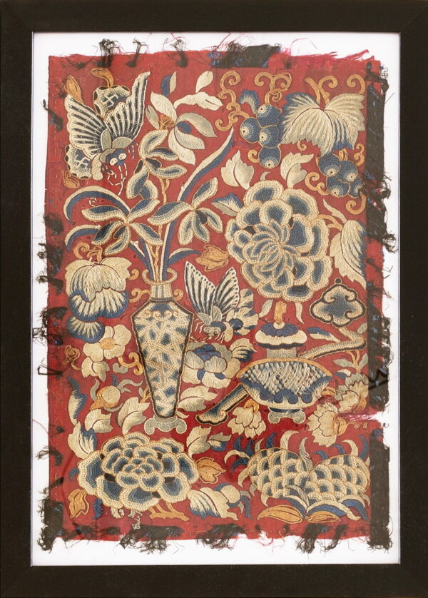 Framed red textile with dense blue and gold embroidery depicting flowers, butterflies, and a patterned vase.