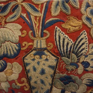 Ornate red textile embroidered with blue, off-white, and metallic gold botanical and vase designs.