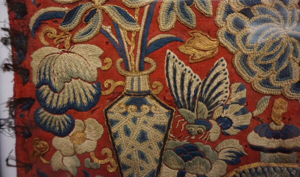 Ornate red textile embroidered with blue, off-white, and metallic gold botanical and vase designs.