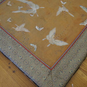 Detail of a gold rug with white bird patterns, an ornate geometric border, resting on brown wooden floors.