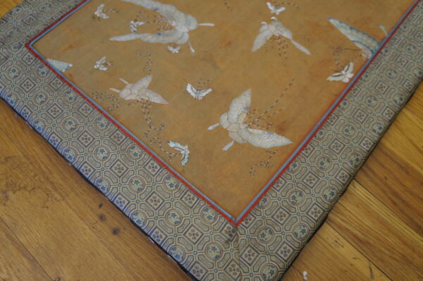 Detail of a gold rug with white bird patterns, an ornate geometric border, resting on brown wooden floors.