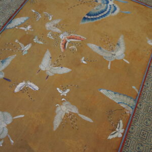 Gold textile rug with blue and white embroidered butterflies and a detailed geometric patterned border rests on a wood floor.