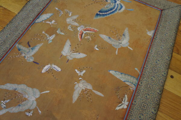 Gold textile rug with blue and white embroidered butterflies and a detailed geometric patterned border rests on a wood floor.