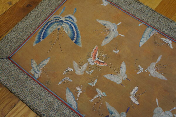 A gold-toned rug features detailed blue and white embroidered butterflies surrounded by a geometric patterned border.