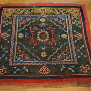 A small square rug with a dark green center, red border, and geometric patterns lies on hardwood flooring.