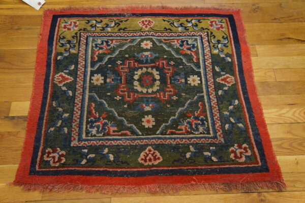 A small square rug with a dark green center, red border, and geometric patterns lies on hardwood flooring.