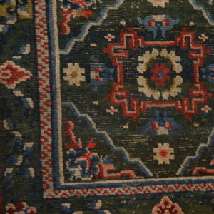 Corner of a multi-colored rug featuring geometric patterns in red, blue, and cream on a dark green field.