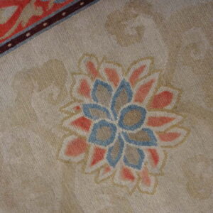 The beige rug field has a central salmon and blue floral medallion and a narrow, dark maroon border stripe.