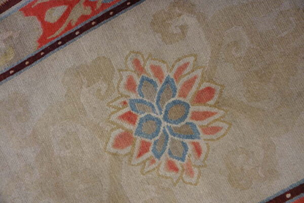 The beige rug field has a central salmon and blue floral medallion and a narrow, dark maroon border stripe.