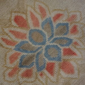 Textured rug close-up displaying a coral and blue stylized floral pattern against a neutral tan ground.