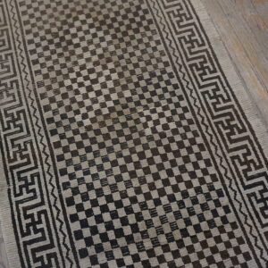A dark brown and beige checkerboard runner rug with geometric borders rests on distressed wood flooring.