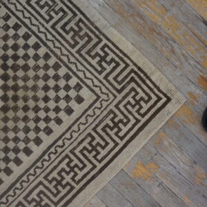 Beige and brown rug with maze-like border and checkered field pattern on weathered gray wood floor.