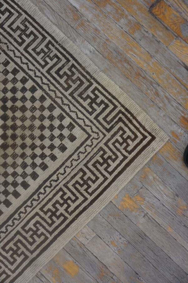 Beige and brown rug with maze-like border and checkered field pattern on weathered gray wood floor.