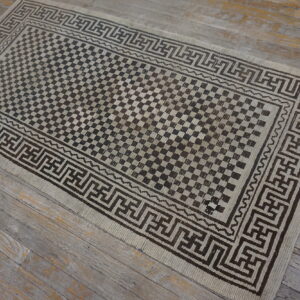 A beige and dark brown geometric rug with a checkered field lies on distressed gray wooden floorboards.