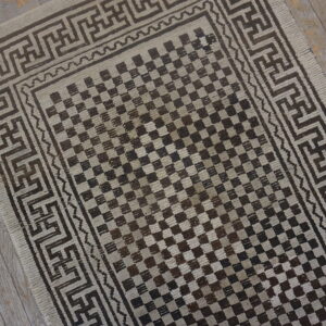 Brown and beige checkerboard rug with geometric meander borders on grey distressed wooden floor.