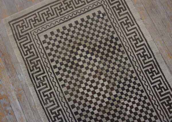 Brown and beige checkerboard rug with geometric meander borders on grey distressed wooden floor.