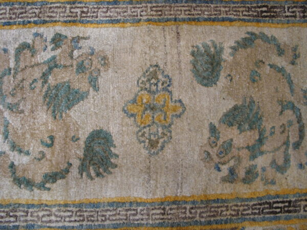 Textured beige rug with faded teal mythological motifs and a detailed blue and yellow geometric border.