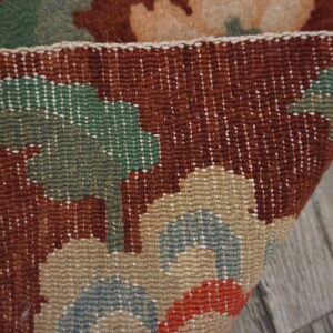 Detail of a rust, green, and tan woven rug folded over gray wood flooring.