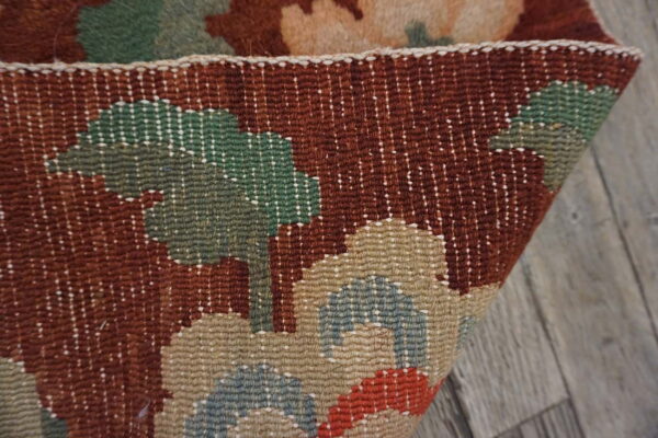 Detail of a rust, green, and tan woven rug folded over gray wood flooring.