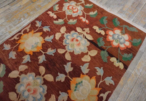 Rust-colored rug with trailing floral designs in cream, blue, and orange sits on distressed wood flooring.
