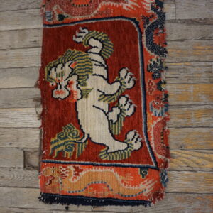 A small high-pile rug fragment featuring a white lion on a red field, lying on distressed gray wood floor.