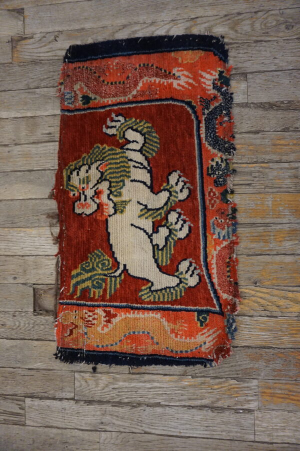 A small high-pile rug fragment featuring a white lion on a red field, lying on distressed gray wood floor.