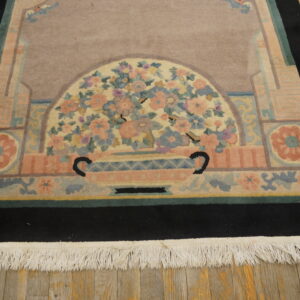 Dusty rose rug with a stylized floral vase design, black border, and visible white fringe on wood planks.