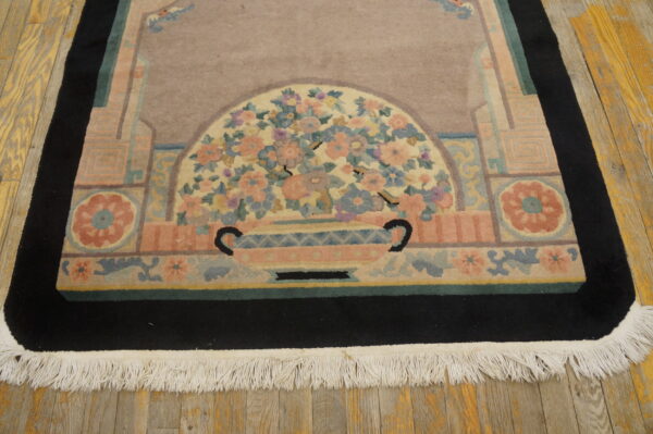Dusty rose rug with a stylized floral vase design, black border, and visible white fringe on wood planks.