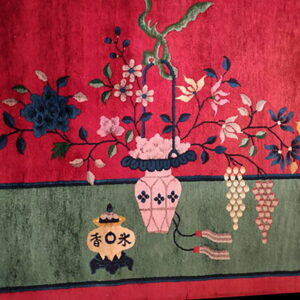 Close-up of a rug featuring a pink vase of multicolored florals on a bright red field above a sage green border.