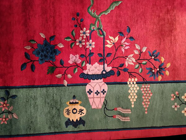 Close-up of a rug featuring a pink vase of multicolored florals on a bright red field above a sage green border.