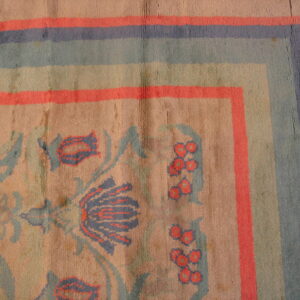 Distressed rug corner with blue and coral striped border surrounding faded floral motifs on a tan ground.