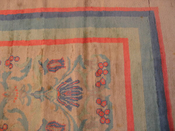 Distressed rug corner with blue and coral striped border surrounding faded floral motifs on a tan ground.