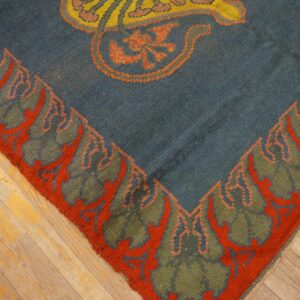 Blue rug corner with olive green and rust border, showing a yellow motif on distressed hardwood.