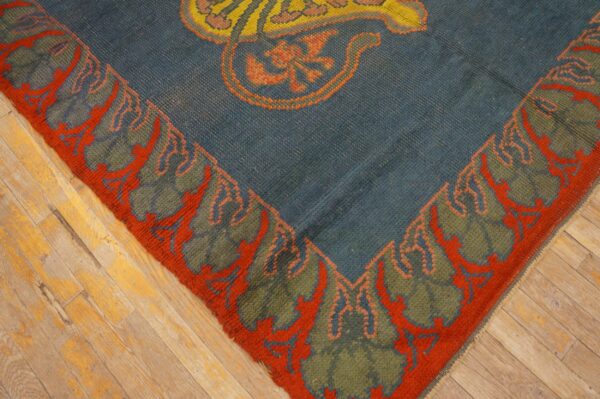 Blue rug corner with olive green and rust border, showing a yellow motif on distressed hardwood.
