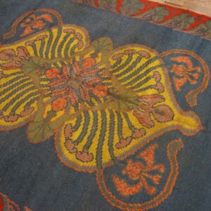 Blue rug with a yellow, striped, and orange medallion, bordered in red and green, resting on wood floor.
