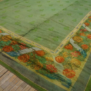 A large, moss green rug with repeating motifs and a wide, multi-colored floral border rests on wood planks.