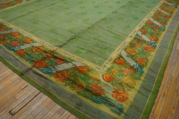 A large, moss green rug with repeating motifs and a wide, multi-colored floral border rests on wood planks.